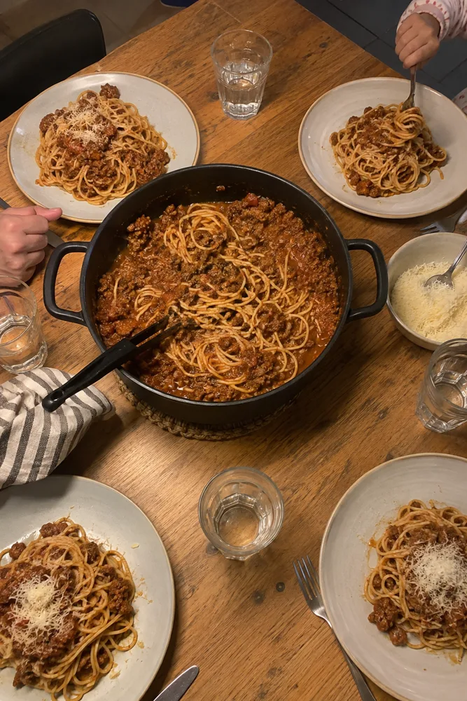 's Avonds aan de keukentafel: een grote pan spaghetti bolognaise, vier borden en een kinderhand met een vork